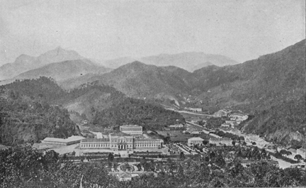 view of the town from a distance. mountains and a large flat wide building in the center.