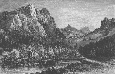 view of a wooded cottage at the foot of a mountain facing the water. there are more mountains and sky in the background.