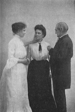 woman in dress touching face of woman in long skirt as a man looks on