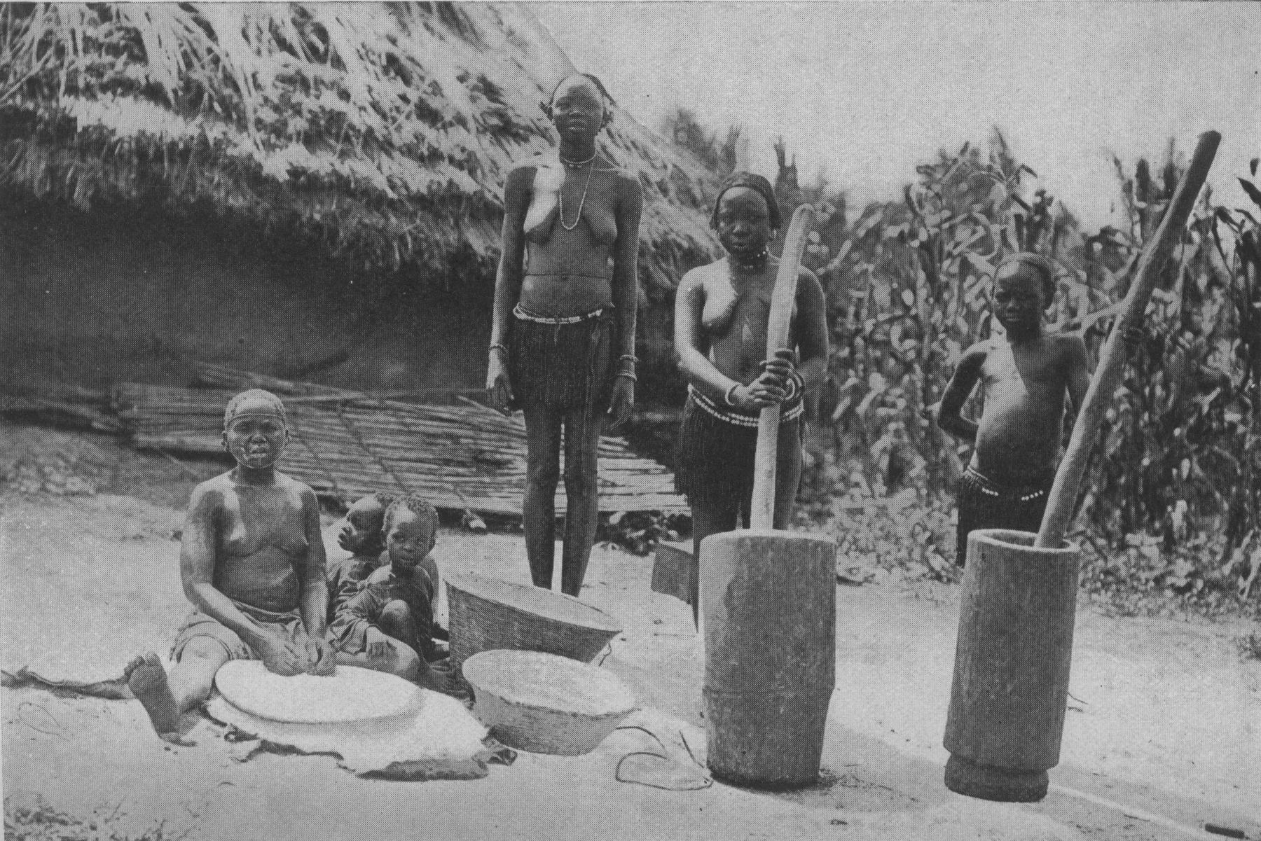 woman and girl using large mortars and pestles with a second woman seated rolling food in a bowl and another woman standing behind