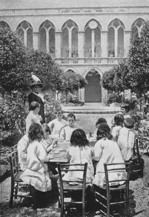 twelve children sitting around a table outside while a woman in hat supervises