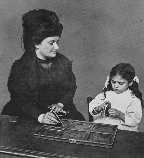 woman pointing at tray with shapes with young girl in pigtails holding circular puzzle piece