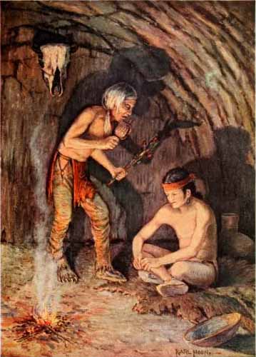 Two Native American men in cave. Young man sits on ground with old man standing before him shaking rattle and prayer-stick.