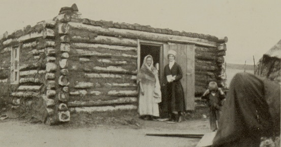 author standing with a woman in front of a one-story log building