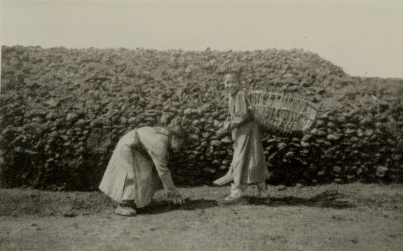 two children standing before a stone wall, one carries a basket on their back