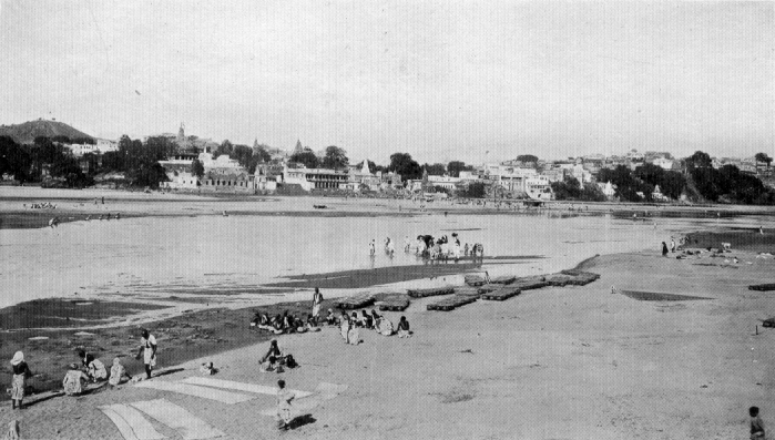 photograph of people on the beach, houses and buildings can be seen across the river.