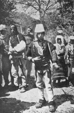 photograph of a group of people, the boy is centrally located and is wearing a cylindrical hat. there are trees in the background and the land looks dry.