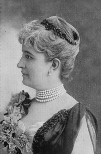 photographic portrait bust profile, her hair is up and she is wearing pearl earrings and necklace with an elaborate beaded dress and sash.
