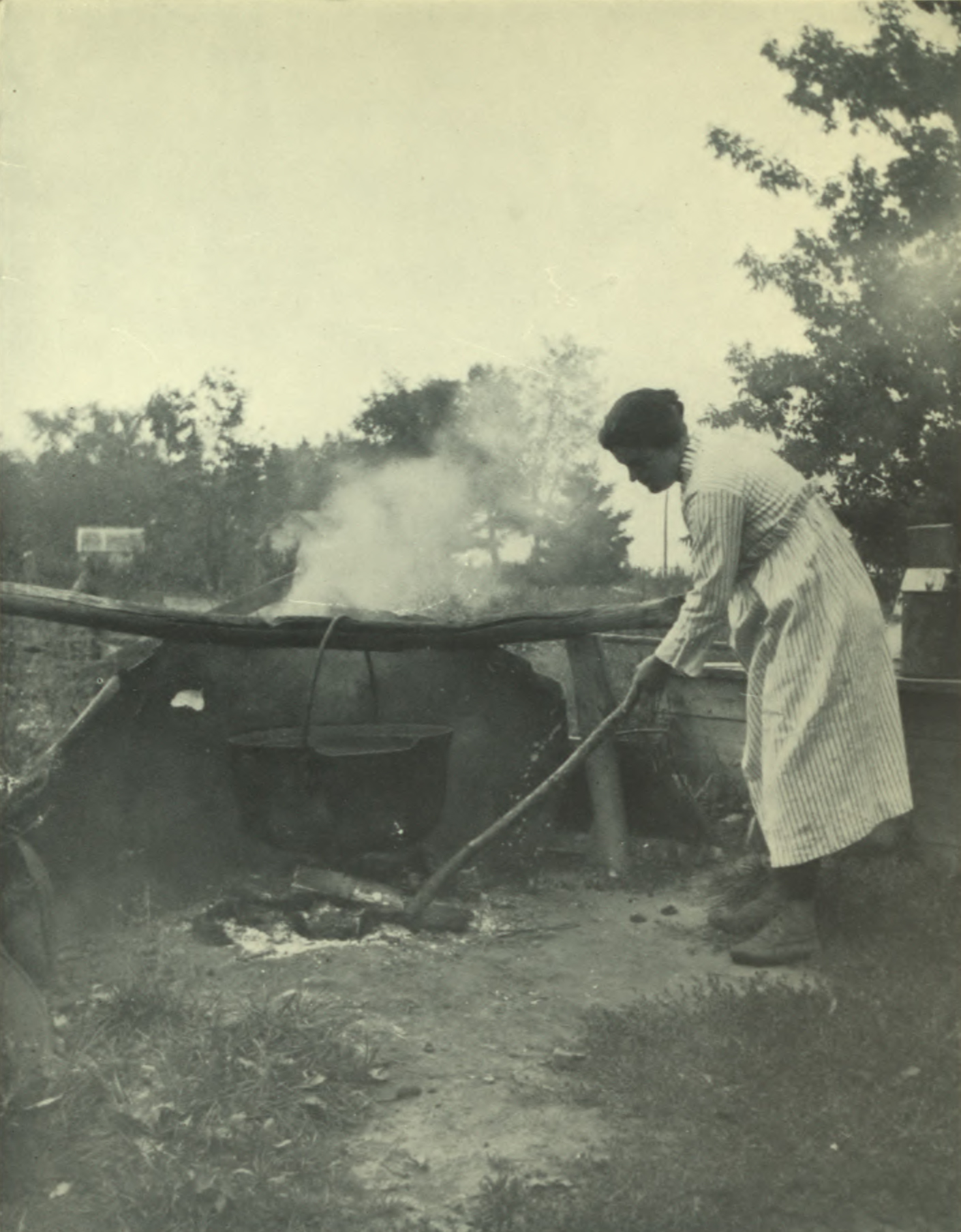 Woman tending to the fire underneath a large caulron handing from a wooden beam.
