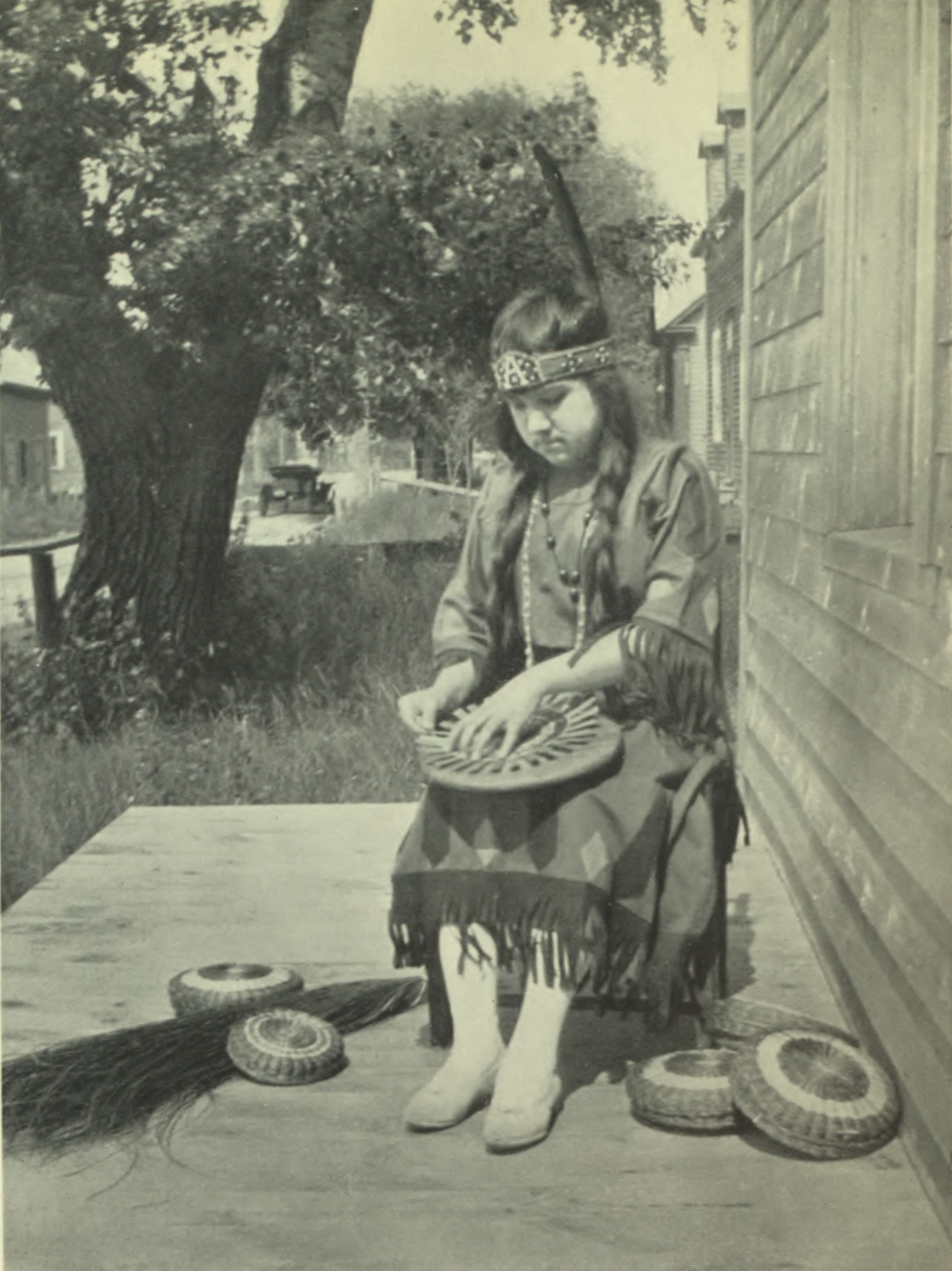 Child in Abenaki traditional clothing sitting outside with some baskets.