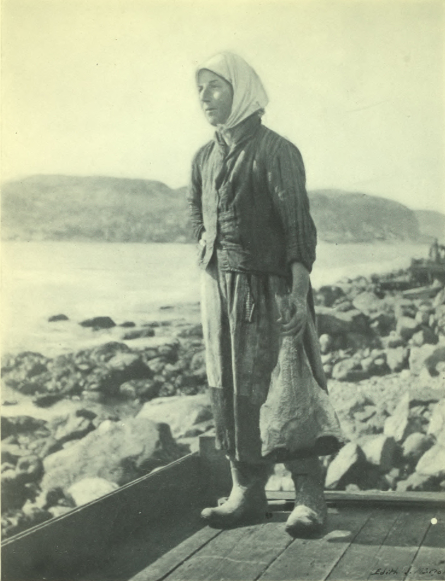 Woman with headscarf, dress, and jacket, standing at the rocky coastline.