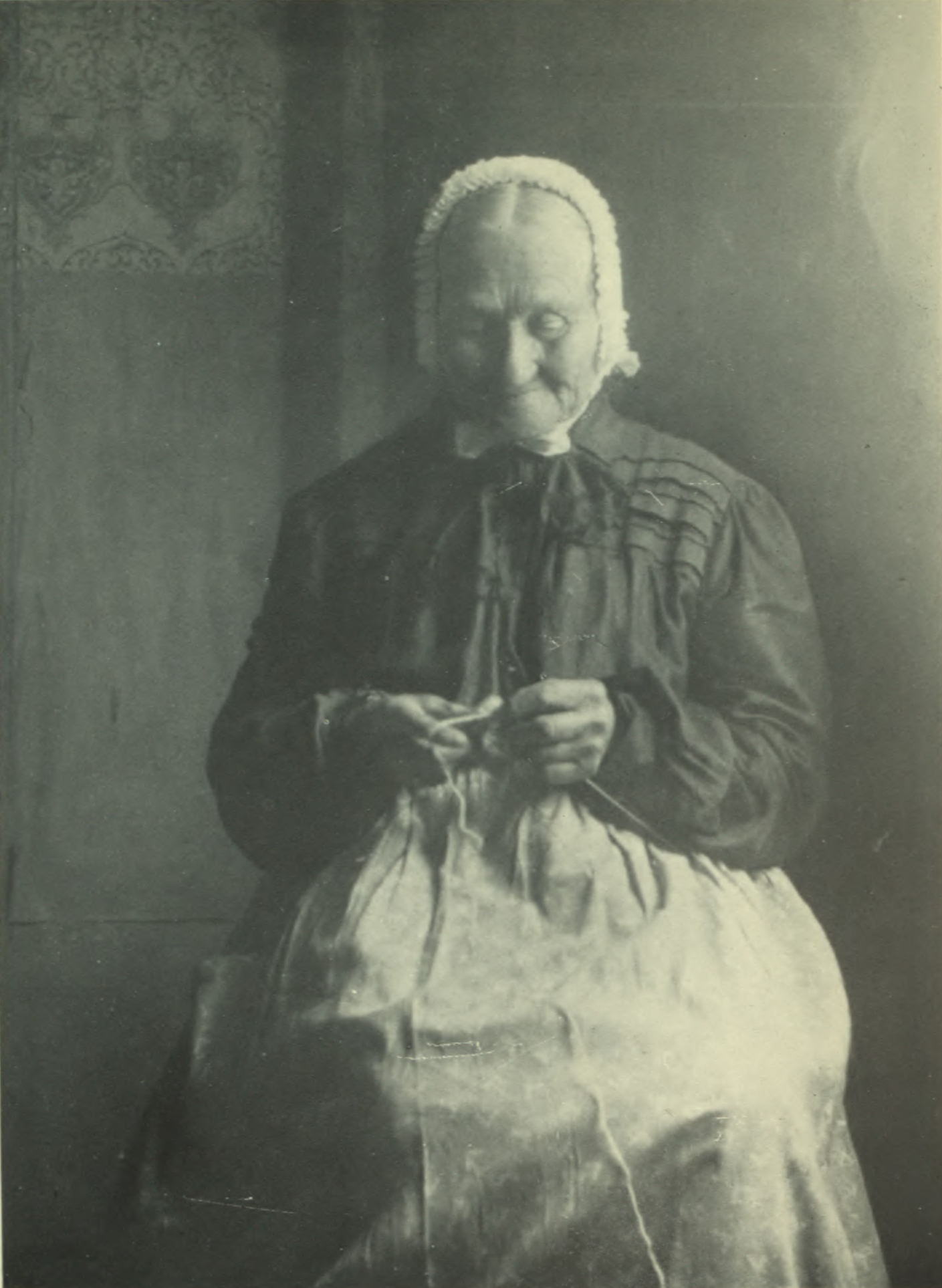 Woman wearing bonnet sewing by the window.