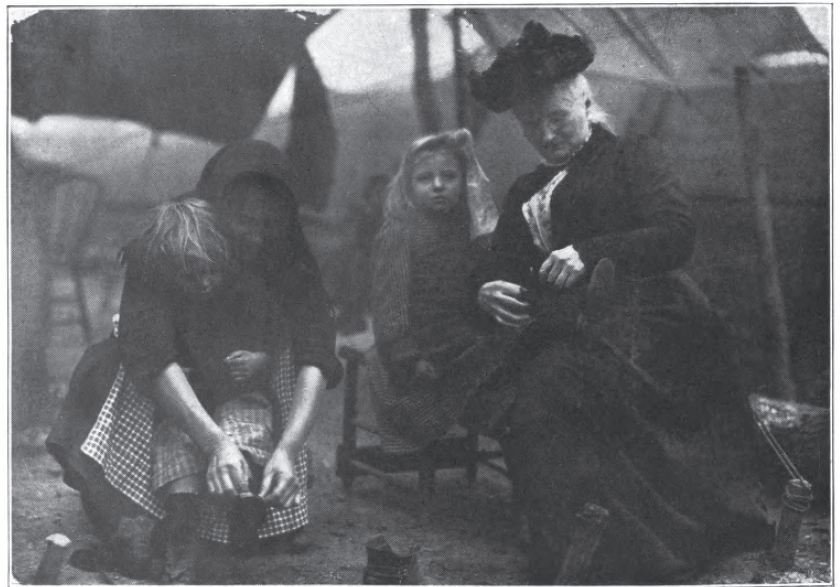 two women, one in apron and one in Victorian dress with hat, tying the shoes of two children