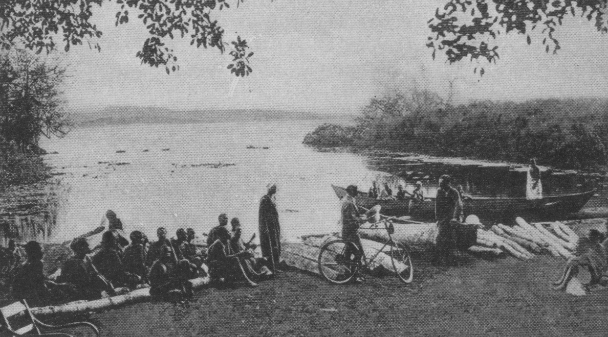 people seated by a lake, one man standing in a boat, another on the shore with a bicycle