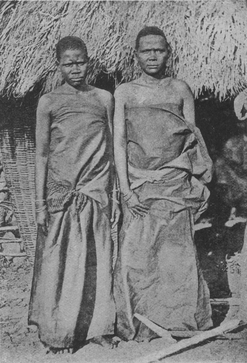 two women in long cloth garments in front of a structure with thatched roof