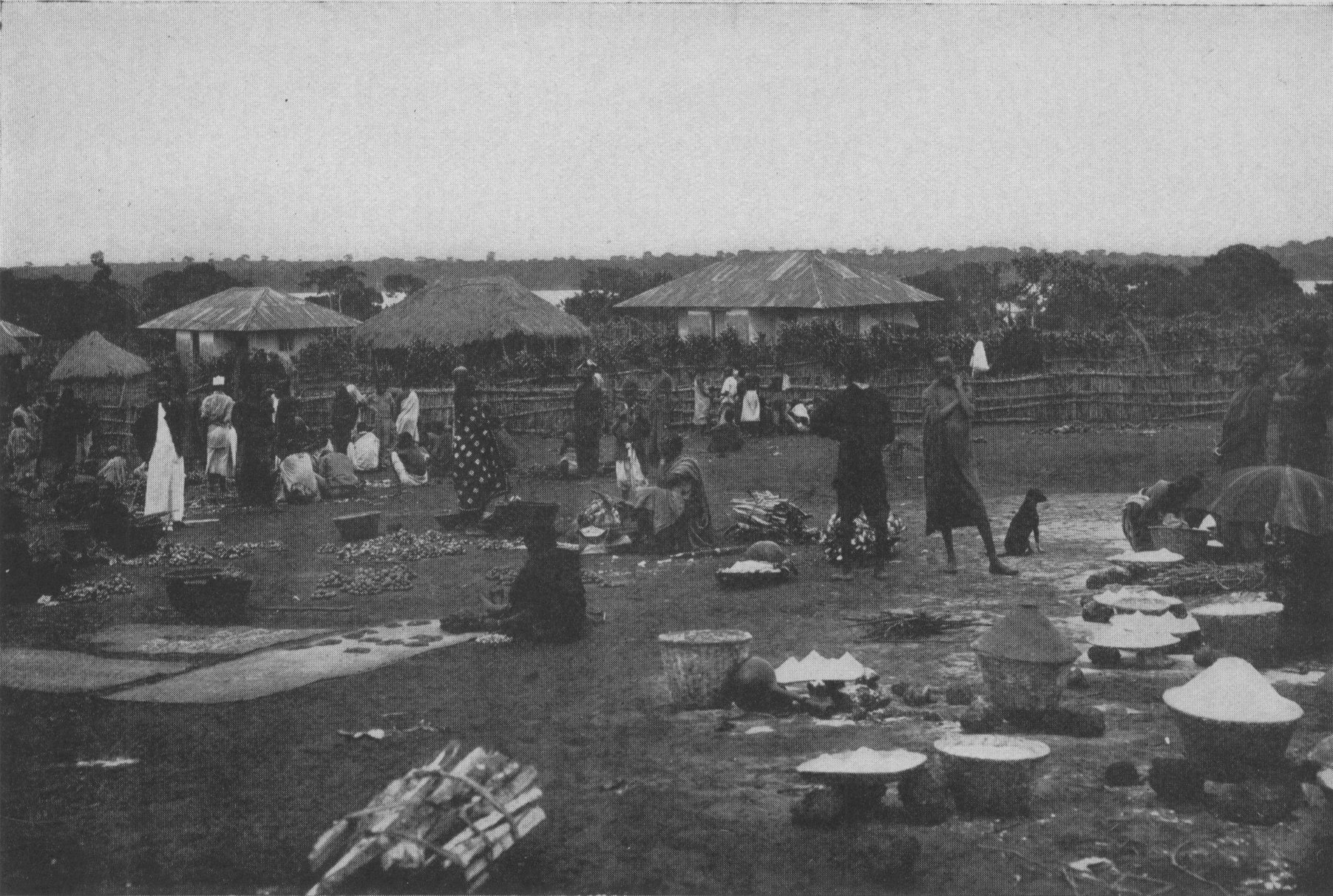 people standing in fenced area with various goods in bowls and low houses in background