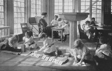 children with blocks and numbered cards with a woman supervising in a room with many windows